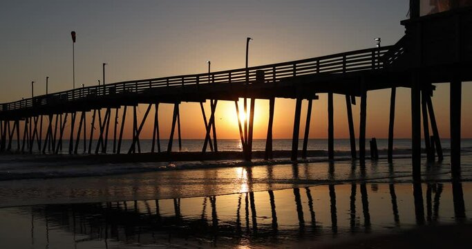 Golden sunrise on the ocean horizon with old wooden fishing pier in silhouette. Light of sunrise shines and reflects on surface of shallow water and wet sand under pier. Peaceful morning beach scene.
