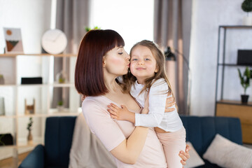 Obraz na płótnie Canvas Lovely family mom and child. Happy affectionate young mother, holds her cute smiling little daughter and kisses her cheek with love, spending free time at home, posing in cozy living room