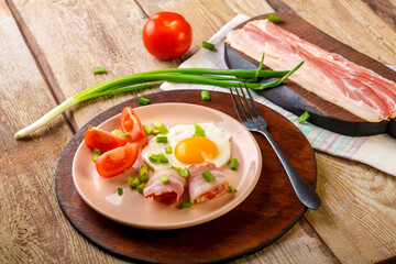 Scrambled eggs with bacon with tomato and green onions and on a wooden table in a plate on a stand with a fork next to chopped bacon and tomatoes.