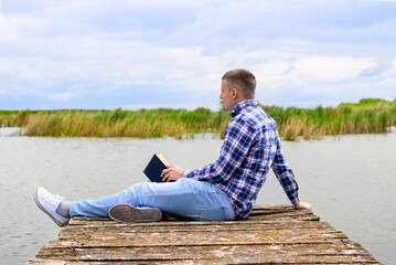 Man sitting on wooden dock at lake. He is holding black book. Nature background. Clouds in the sky.