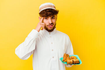 Young Moroccan man wearing the typical arabic costume eating Arabian sweets isolated on yellow background showing a disappointment gesture with forefinger.