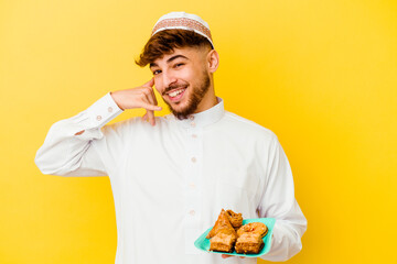 Young Moroccan man wearing the typical arabic costume eating Arabian sweets isolated on yellow background showing a mobile phone call gesture with fingers.
