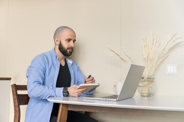 A guy in a blue shirt and black jeans is studying online from home sitting at a desk using a computer and a diary for writing