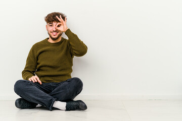 Young Moroccan man sitting on the floor isolated on white background excited keeping ok gesture on eye.