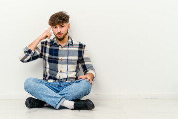 Young Moroccan man sitting on the floor isolated on white background pointing temple with finger, thinking, focused on a task.