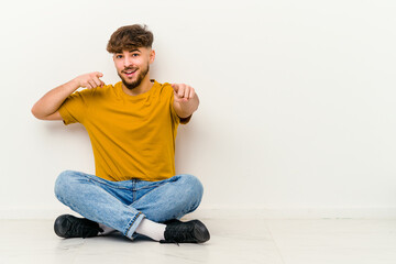 Young Moroccan man sitting on the floor isolated on white background cheerful smiles pointing to front.