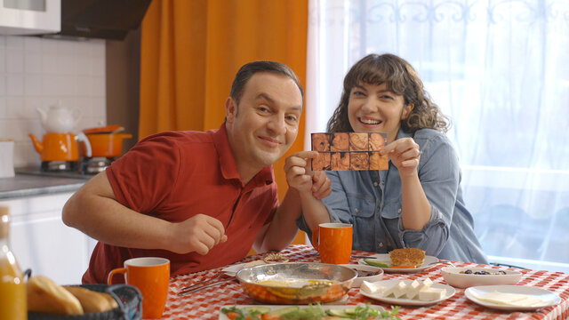 Young couple having a video call with their loved ones while having breakfast at home. They give good news by showing an ultrasound photo during the conversation. Front camera view.