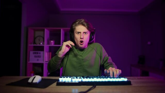 Portrait of a surprised young male gamer in casual clothes and headset sitting in a room with red light at night and looking at the camera with a shocked face and shouts WHAT