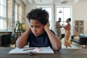 Mixed-race annoyed schoolboy covering ears by hands while sitting by table and trying to do homework