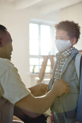 Young father buttoning up jacket of his little son with protective mask on face and backpack on back