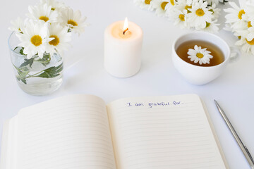 Close up of handwritten text "I am grateful for..." in foreground with notebook, pen, cup of tea, flowers