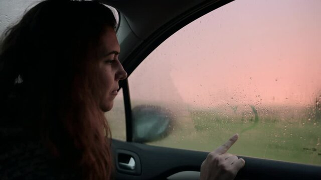 Side view young woman drawing a heart on the fogged window glass inside a car during sunset with rain.