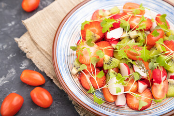 Vegetarian fruits and vegetables salad of strawberry, kiwi, tomatoes, microgreen sprouts on black concrete background. Side view, selective focus.