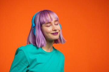 Exceptional woman with dyed violet hair listening music in headphones and singing on orange...