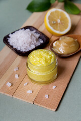 Natural cosmetic scrub with sea salt, honey and lemon in a glass jar on a deoevyan stand, next to half a lemon, a wooden spoon with honey, a jar of sea salt and pieces nearby, vertical photo; close-up