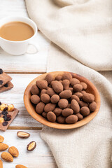 Almond in chocolate dragees in wooden plate and a cup of coffee on white wooden background. Side view.