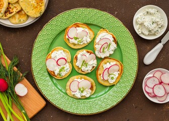 Snack mini pancakes with a spread of salted cottage cheese, herbs and slices of radish on a green plate on a brown concrete background.