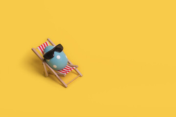 Creative funny idea with Easter egg with sunglasses while sitting on deck chair on illuminating yellow background.