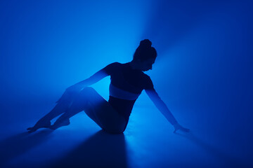 Naklejka na ściany i meble Professional ballerina dancing ballet in spotlights smoke. Beautiful young female wearing black bodysuit on floodlights background.