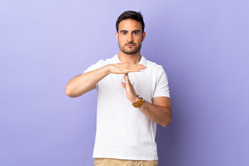Young handsome man isolated on purple background making time out gesture