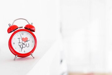 a red retro alarm clock with a red heart and the inscription on the dial I love you stands on a white shelf in a white room