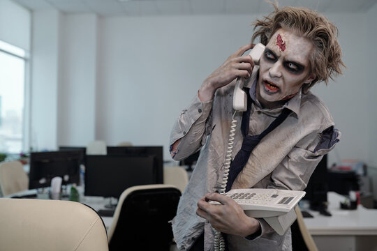 Zombie businessman with phone receiver by ear standing by workplace in front of camera