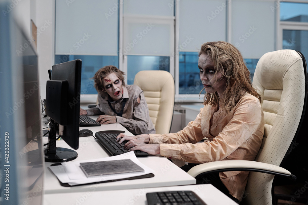 Poster young spooky zombie businesswoman sitting by desk in front of computer monitor and networking