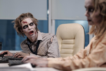 Zombie businessman with greasepaint on face and hands sitting by workplace and looking at co-worker