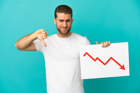 Handsome blonde man over isolated blue background holding a sign with a decreasing statistics arrow symbol and doing bad signal