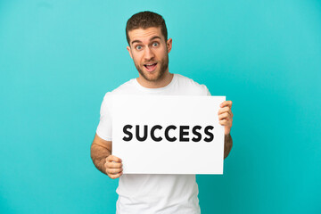 Handsome blonde man over isolated blue background holding a placard with text SUCCESS with surprised expression