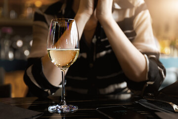 Closeup view of white wine glass on table against young adult woman sitting on ddating at cafe or restaurant at warm sunset time. Alone single female person drinking champagne at bar