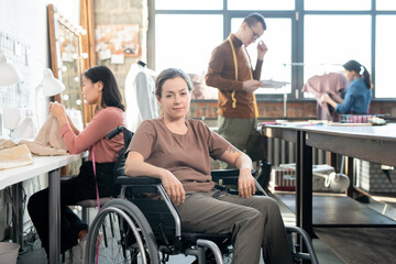 Fototapeta na wymiar Young smiling seamstress in casualwear sitting in wheelchair against group of working colleagues
