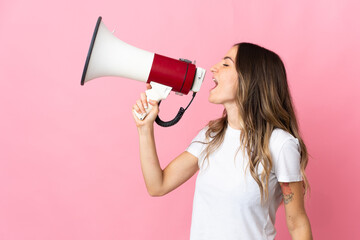 Young Romanian woman isolated on pink background shouting through a megaphone to announce something in lateral position