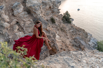 A girl with loose hair in a red dress sits on a rock rock above the sea. In the background, the sea. The concept of travel.