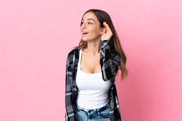 Young Romanian woman isolated on pink background listening to something by putting hand on the ear