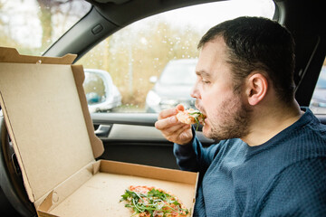 Man with beard eats pizza in his cart. Eat takeaway food in the car. Due to closed restaurants during the pandemic, you are forced to order food. Flatbread in a cardboard box that says 