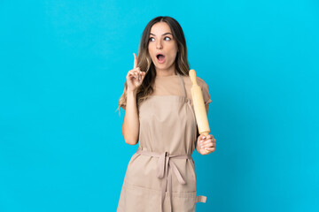 Young Romanian woman holding a rolling pin isolated on blue background thinking an idea pointing the finger up