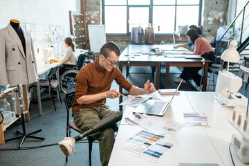 Young serious designer with prosthetic leg sitting by desk in front of laptop while making sketches