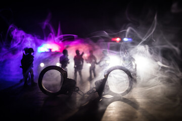 Police raid at night and you are under arrest concept. Silhouette of handcuffs with police car on backside. Image with the flashing red and blue police lights at foggy background.