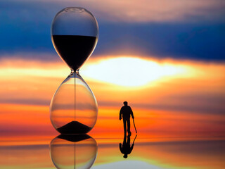 miniature people. silhouette of an elderly man walking towards the sunset next to an hourglass. end of life. - obrazy, fototapety, plakaty