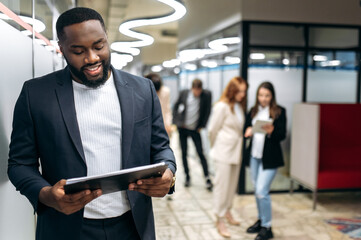 Fototapeta na wymiar Influential businessman in stylish formal suit is looking at the tablet, smiling, browsing internet. Successful african american male employee is standing in office hall, working on project
