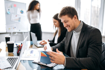 Joyful male employee doing paperwork on business meeting, smiling. Successful young manager in formal suit learning financial graphs, working with collegues on corporate project