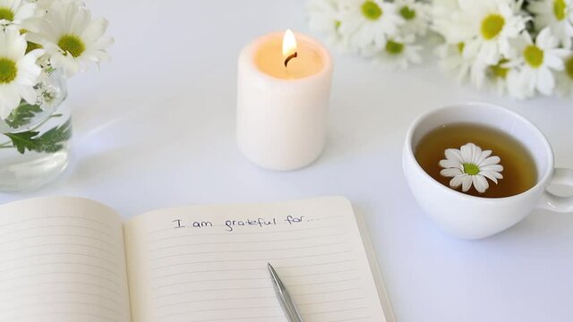 Close up of handwritten text "I am grateful for..." in foreground with notebook, pen, cup of tea, flowers