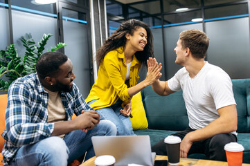 Satisfied multiracial students or colleagues sitting in modern office, well done with corporate project. Successful coworkers giving high five to each other, smiling, happy about good work