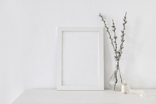 Easter spring still life. Blank vertical picture frame mockup on white wooden table. Blooming pussy willow branches with catkins in glass vase. Art concept. Scandinavian interior design, no people.