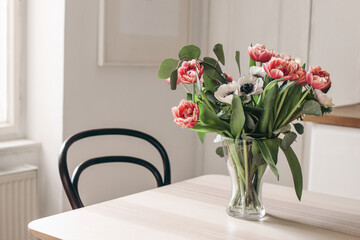 Spring flowers in glass vase on wooden table. Blurred kitchen background with old chair. Bouquet of red tulips, white anemone flowers and eucalyptus branches. Contemporary elegant, Scandinavian