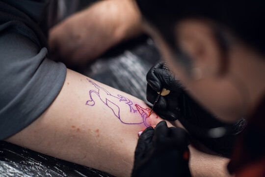 Close-up photo of a girl tattoo artist putting a tattoo on the arm, of a young guy in a tattoo parlor using the handpoke method
