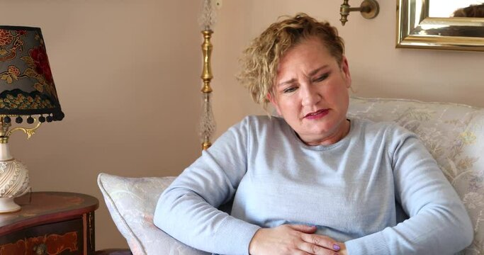 Portrait of a woman with blonde hair sitting on sofa having strong stomach ache  hands on abdomen