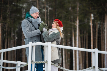 A young husband and wife stand on the bridge in an embrace against the background of a winter forest.