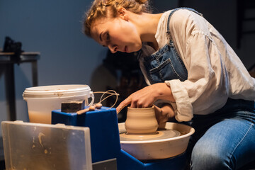 Fototapeta na wymiar a young girl in a white blouse and denim overalls makes a pot on a potter's wheel with her hands